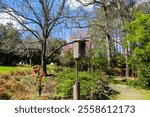 a small brown wooden bird house on a post surrounded by pink trees, lush green trees and plants, bare winter trees with green grass, blue sky and clouds at Smith-Gilbert Gardens in Kennesaw Georgia	