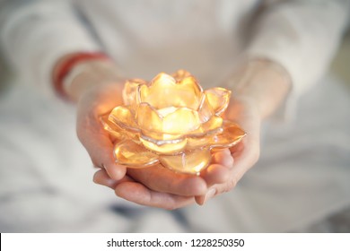 Small burning candle in an transparent orange lotus shaped glass candle holder laying on streched woman's hands with  soft-focused white women clothes in the background