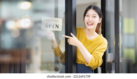 Small Business Concept. Smiling Asian Woman Cafe Stuff In An Apron Points Finger On An Open Sign Board In Her Hand. A Multiracial Cafe Owner Invites To Visit Reopened Location