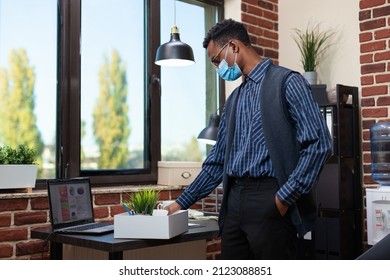 Small Business Employee Wearing Covid Mask Gathering Personal Belongings In Tray Going To Work From Home Because Of Covid 19 Pandemic. Startup Worker Being Fired Standing Next To His Former Office.