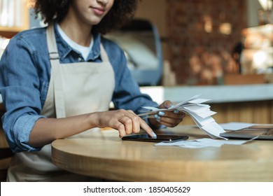 Small Business Management And Accounting. Busy African American Young Lady Owner In Apron Works With Papers, Counts Costs And Profits In Interior Of Modern Cafe, Cropped, Close Up, Copy Space