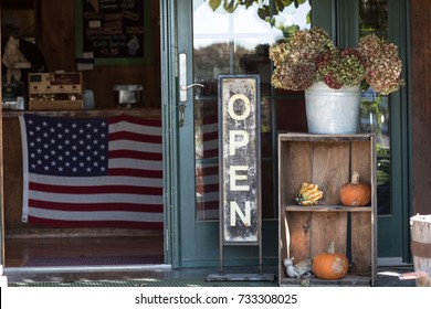 A Small Business Is Open For Business With A Welcoming Open Sign, Pumpkins And Dried Flowers. Inside The Store Hangs An American Flag. 