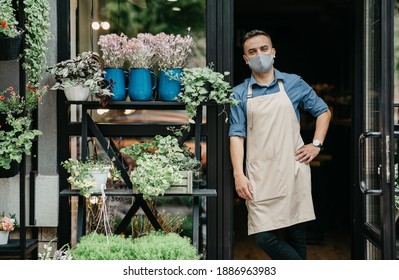 Small business owner and flower startup during covid-19 quarantine. Attractive smiling young guy in apron and protective mask stands at front door and waits for clients in studio with pots of plants - Powered by Shutterstock