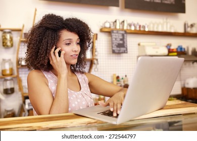 Small Business Owner In Her Coffee Shop Typing On Her Laptop While Talking On The Phone