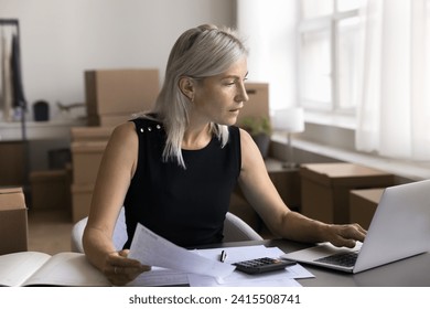 Small business owner, individual entrepreneur serious businesswoman check invoices, counts profit, pay bills, manage own company finances, sit at desk using laptop, doing paperwork in warehouse office - Powered by Shutterstock