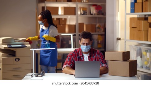 Small Business Owner In Mask Working On Laptop In Warehouse With Maid Cleaning On Background. Female Janitor Disinfect Furniture With Detergent Spray In Small Storehouse