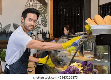 Small business owner multitask lifestyle concept, Asian coffee shop owner man or worker cleaning bakery cabinet for opening, multitask of entrepreneur wearing apron looking camera while working - Powered by Shutterstock