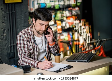 Small Business Owner On Phone And Computer In Store. Sport Shop Worker Making Call With Cell Phone. Salesman Working And Talking On Cell Phone In Bicycle Shop. Seller Taking Orders By Phone.