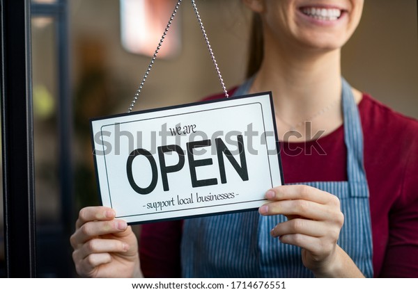 Small business owner smiling while turning the\
sign for the reopening of the place after the quarantine due to\
covid-19. Close up of woman’s hands holding sign now we are open\
support local business.