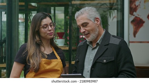 Small Business Owner Speaking With Female Employee Barista Wearing Apron. Boss Manager Talking With Staff Explaining Restaurant Business Holding Tablet Device