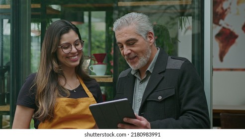 Small Business Owner Speaking With Female Employee Barista Wearing Apron. Boss Manager Talking With Staff Explaining Restaurant Business Holding Tablet Device