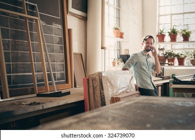 Small Business Owner Talking On His Phone While Smiling And Standing Casually In His Studio Workshop