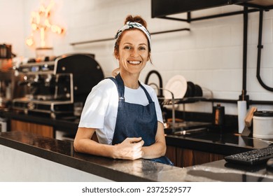 Small business owner wearing an apron stands behind the counter of her bustling coffee bar. Woman managing orders with a smile, ensuring that every customer receives a delicious cup of coffee.