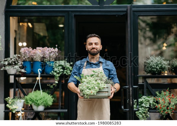 Small business owner works in flower shop and\
customer service. Confident smiling young bearded european male in\
apron carries wooden box with plants, on shop or eco cafe\
background, empty space