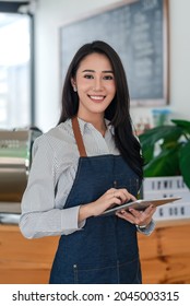 Small Business Owner Of Young Asian Woman Wearing Aprons Holding A Tablet At A Café. Looking At The Camera.