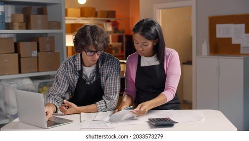 Small Business Owners Calculate Finances In Warehouse. Diverse Male And Female Managers Using Laptop And Calculator Paying Bills And Count Profit