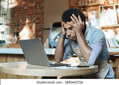 Small Business Owners, Coronacrisis And Problem During Covid-19 Quarantine. Sad Young African American Man Holds His Head And Looks On Laptop Trying To Avoid Bankruptcy In Interior Of Modern Loft Cafe