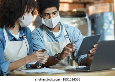 Small Business Owners At Social Distancing Of Covid Quarantine. Busy African American Couple In Aprons And Protective Masks Work With Laptop And Tablet To Attract Clients In Trendy Cafe Or Restaurant