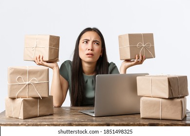 Small Business Owners, Startup And E-commerce Concept. Confused Asian Female Entrepreneur Working With Laptop, Packing Items For Shipping, Puzzled As Holding Boxes, Mixed Up Orders