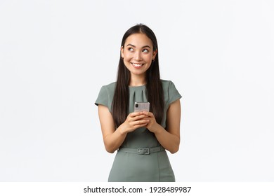 Small business owners, women entrepreneurs concept. Dreamy smiling stylish asian blogger, looking upper left corner thoughtful, creating content, thinking while texting message, using smartphone - Powered by Shutterstock