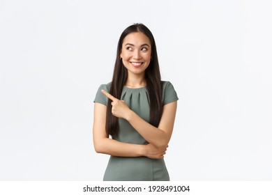 Small Business Owners, Women Entrepreneurs Concept. Intrigued Happy Asian Female In Dress Found Excellent Choice, Pointing Finger Left And Looking Interested, Smiling Satisfied, White Background