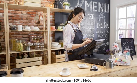 Small Business People And Service Concept. Young Asian Girl Waitress At Wood Bar Counter In Cafe Store. Woman Bartender In Apron Using Pos Sale Terminal Machine To Enter Customer Order From Note.