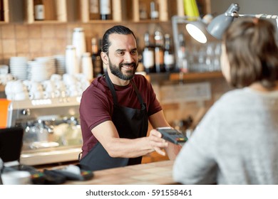 Small Business, People And Service Concept - Happy Man Or Waiter In Apron With Card Reader And Customer Paying At Bar Of Coffee Shop