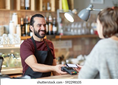 Small Business, People And Service Concept - Happy Man Or Waiter In Apron With Card Reader And Customer With Smartphone Paying At Bar Of Coffee Shop