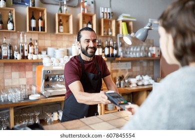 Small Business, People And Service Concept - Happy Man Or Waiter In Apron With Card Reader And Customer Paying At Bar Of Coffee Shop