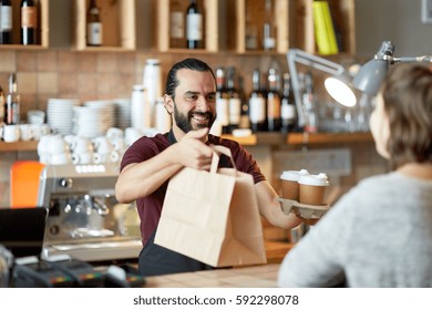 small business, people, takeaway and service concept - happy man or waiter giving bag and paper cups with hot drinks to customer at coffee shop