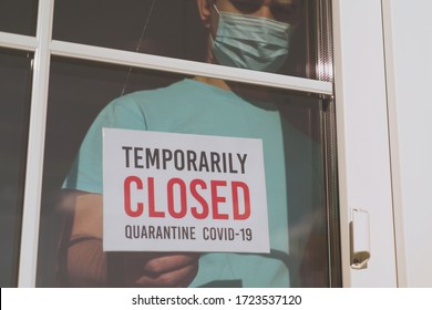 Small Business Sign On The Storefront Window Closed Because Of Coronavirus Or COVID19. Business Owner In Medical Mask Puts A CLOSED Sign On The Front Door Due To The Coronavirus COVID19 Pandemic.