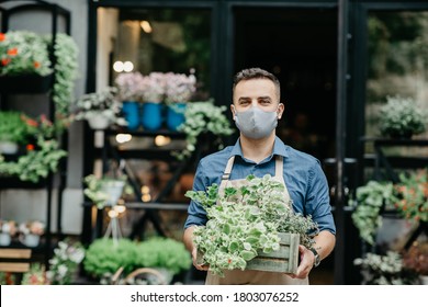 Small Business And Start Of Working Day. Man In Protective Mask Takes Out Box Of Plants Outside In Front Of Flower Shop