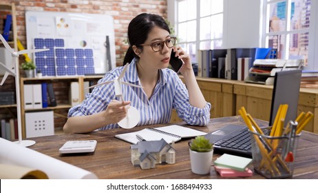 Small Business Startup And People Concept. Creative Female Engineer Worker With Laptop Computer Calling On Smartphone. Young Lady Architect Discussing On Cellphone With Green Energy Building Plan.