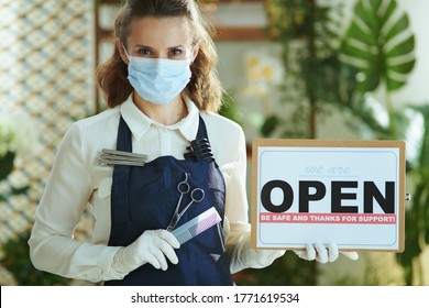 Small business survival after covid-19 pandemic. Portrait of elegant small business owner woman in apron with medical mask, gloves, hair comb and scissors showing open sign. - Powered by Shutterstock