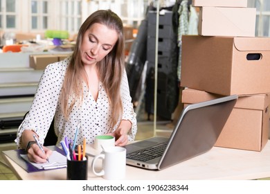 Small Business, Young Female Entrepreneur Working At A Desk, Stack Of Cardboard Boxes Ready For Delivery