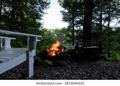 A small campfire burns in a stone ring beside a white wooden bench and stacked firewood at a wooded clearing in the quiet blue light of dusk.