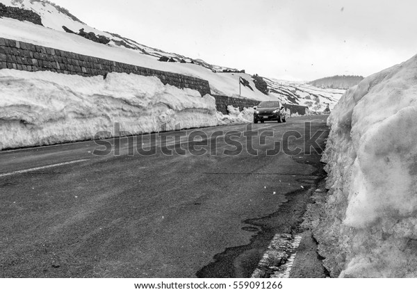 A small car runs along a stretch of\
road in the mountains while it snows and the edge of the road is\
covered by a high layer of snow, in black and\
white