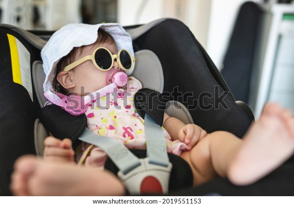 Small caucasian baby four months old sitting\
in the car seat at home in room with eyeglasses and hat on her head\
ready for travel to go out on\
vacation