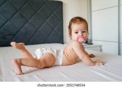 Small Caucasian Baby Girl Six Months Old Lying On The Bed Naked Belly Down With Nipple Pacifier Wearing Diaper At Home In Bright Room Copy Space Side View