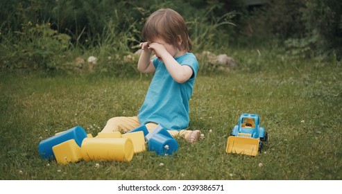 Small Caucasian Child Is Sitting On The Grass And Crying Loudly. There Are Various Toys Around The Child. The Concept Of A Sharp Change Of Mood In Young Children