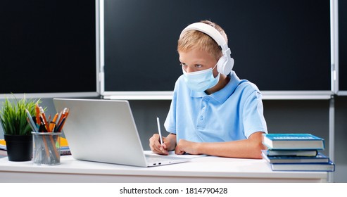 Small Caucasian Teen Boy In Medical Mask And Headphones Sitting At Desk In School. Teenage Schoolboy Doing Homework And Writing Exercise In Copybook In Front Of Laptop Computer. Covid-19 Concept.