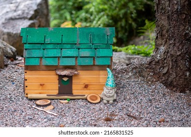 A small ceramic garden gnome with a green hat stands next to a tiny colorful fantasy house with a door and a slanted roof. The magical wooden building is among green garden shrubs and base of a tree.