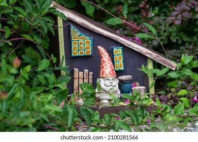 A small ceramic garden gnome with a red hat stands next to a tiny colorful fantasy house with windows, a door, and a slanted roof. The magical wooden building is among green garden shrubs and plants. 