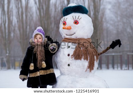 Small cheerful girl near big funny snowman. Cute little girl has fun in winter park, wintertime