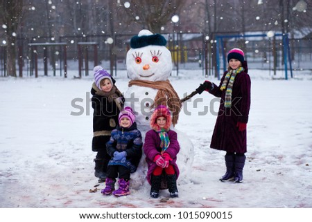 A small cheerful girls near big funny snowman. A cute little sisters has fun in winter park, wintertime