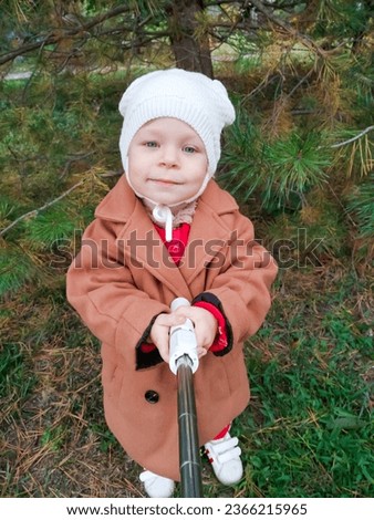 A small child in an autumn coat smiles at the camera and takes a selfie stick. Girl in park in autumn taking selfie photo. Fall and leisure concept. Smile portrait boy autumn jacket , fun emotion.