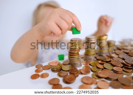 small child, blonde girl 3 years old playing with cash, house model on stacks euro currency coins, pocket money, concept home mortgage, home insurance, financial literacy of children, personal savings