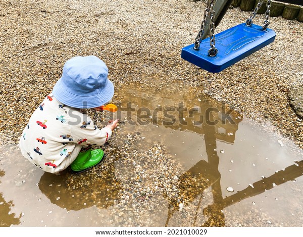 Small child in blue hat and green Wellington boots\
playing in the splash with gravel stones after rain. Kid’s\
playground, being dirty, blue\
swing.