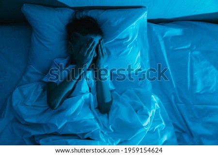 Small child boy lies in bed in dark night and covers his face with his hands in fear, afraid of nightmares and terrible dreams in children. Concept of horror and domestic violence