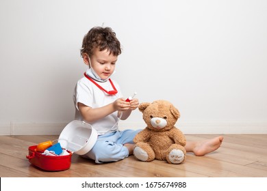 A Small Child, A Boy In A Medical Mask With A Stethoscope On His Neck Plays A Doctor With A Children's Medicine Cabinet, Gives A Teddy Bear An Injection And Measures The Temperature.